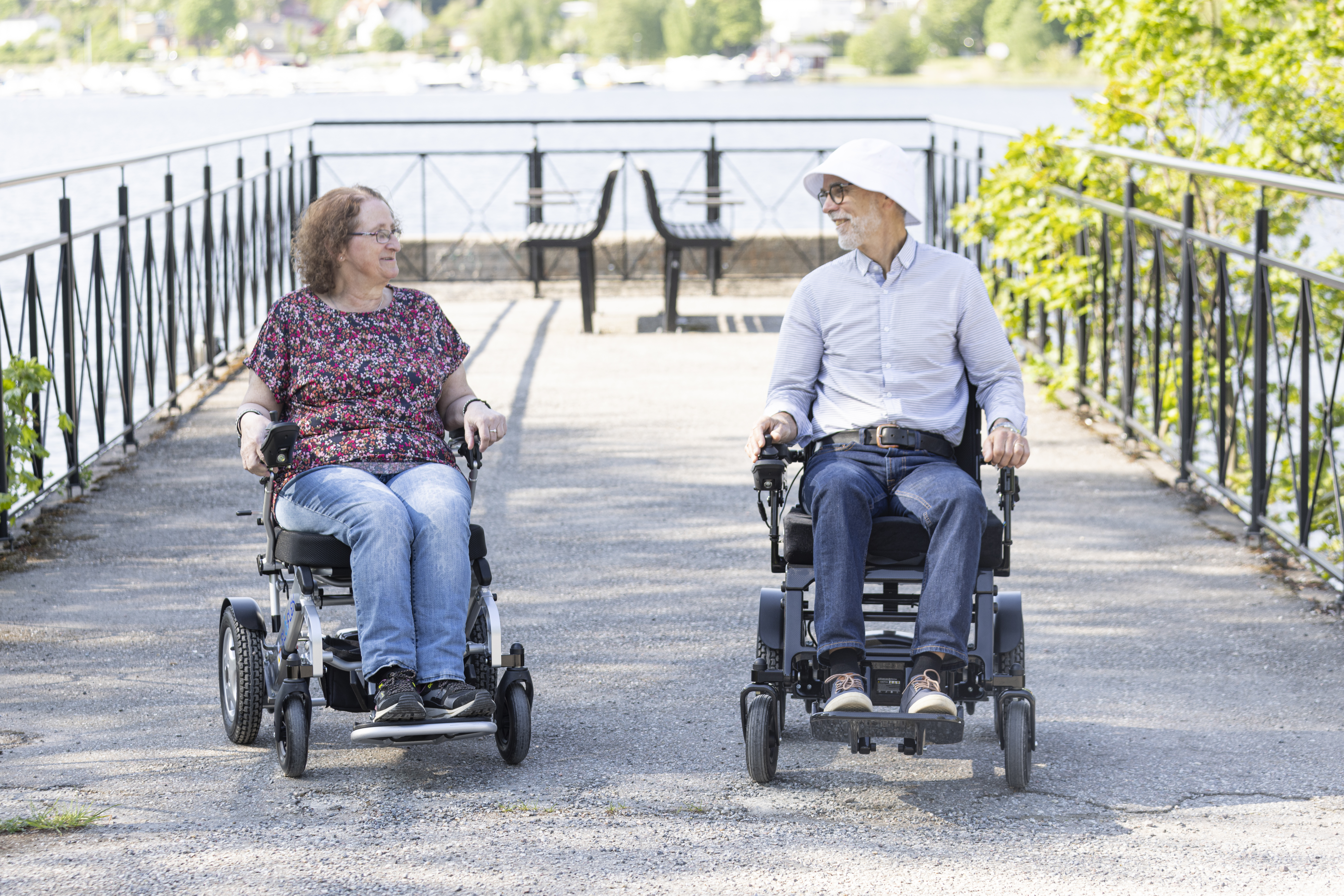 To eldre personer i Eloflex elektriske rullestoler kjører side om side på en gangvei ved vannet og smiler til hverandre.
